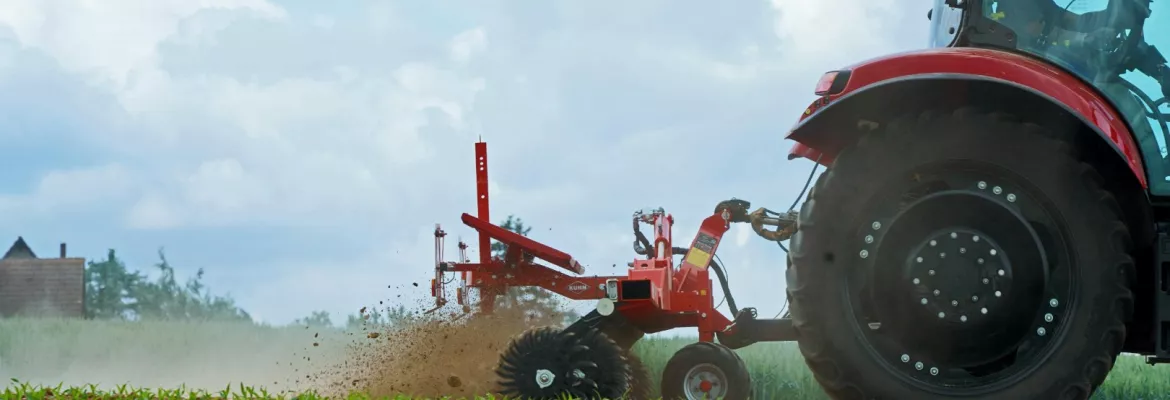 Un tracteur rouge tracte la houe rotative Starliner à travers un champ de terre, soulevant un nuage de poussière. Des cultures vertes poussent en rangs à proximité, sous un ciel nuageux, tandis que des bâtiments agricoles sont visibles à l’arrière-plan.