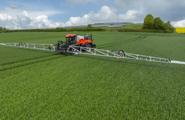 Un tracteur rouge équipé du pulvérisateur automoteur ARTEC F40 EVO de KUHN réalise une opération de pulvérisation dans un champ de culture verdoyant sous un ciel nuageux, avec des panneaux solaires visibles sur les collines à l’arrière-plan.