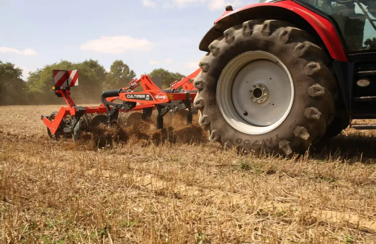 Tracteur agricole équipé d'un cultivateur à dents KUHN Cultimer réalisant un déchaumage dans un champ après récolte, avec dégagement de poussière lors de la préparation du sol.