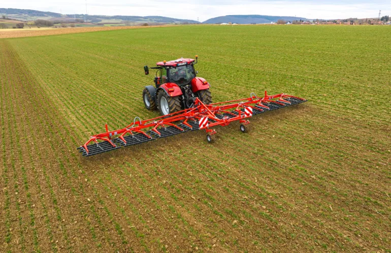 Un tracteur rouge tracte la herse étrille TINELINER de KUHN dans un champ verdoyant aux rangs de culture bien visibles, entouré de collines ondulantes et de terres agricoles à l'horizon sous un ciel