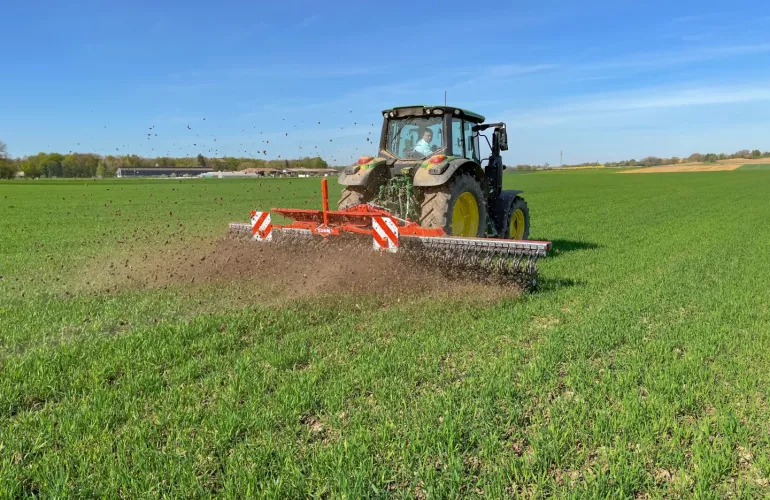 Un tracteur tracte la houe rotative STARLINER rouge de KUHN dans un champ verdoyant, travaillant le sol en bandes. La scène illustre un chantier agricole sous un ciel bleu dégagé, avec des oiseaux visibles dans les airs.