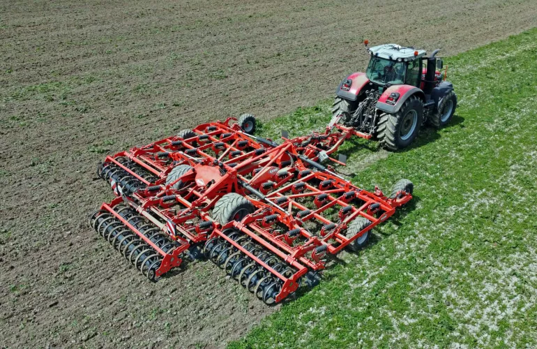Un tracteur rouge et gris tracte le cultivateur à dents Highlander de KUHN, équipé de plusieurs rangées de disques, à travers un champ agricole brun, tandis que des rangs de cultures verts sont visibles à proximité de la zone travaillée.