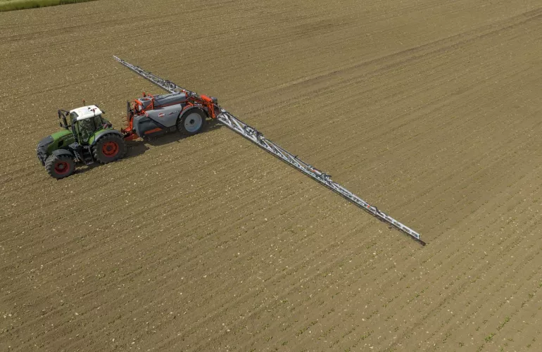 Un tracteur vert tractant le pulvérisateur traîné KARAN intervient dans une parcelle agricole au sol brun marqué par des sillons visibles, assurant une pulvérisation efficace dans les cultures.