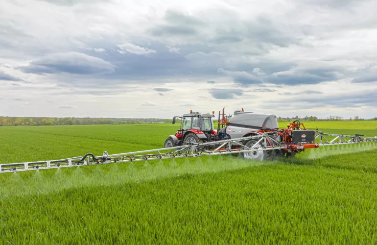 Un tracteur rouge équipé du pulvérisateur traîné LEXIS réalise une opération de pulvérisation dans un champ d’un vert intense sous un ciel nuageux, avec des arbres visibles à l’horizon.
