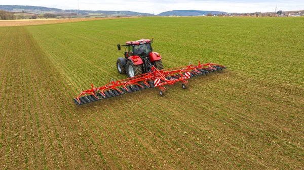 Un tracteur rouge tracte la herse étrille TINELINER de KUHN dans un champ verdoyant aux rangs de culture bien visibles, entouré de collines ondulantes et de terres agricoles à l'horizon sous un ciel