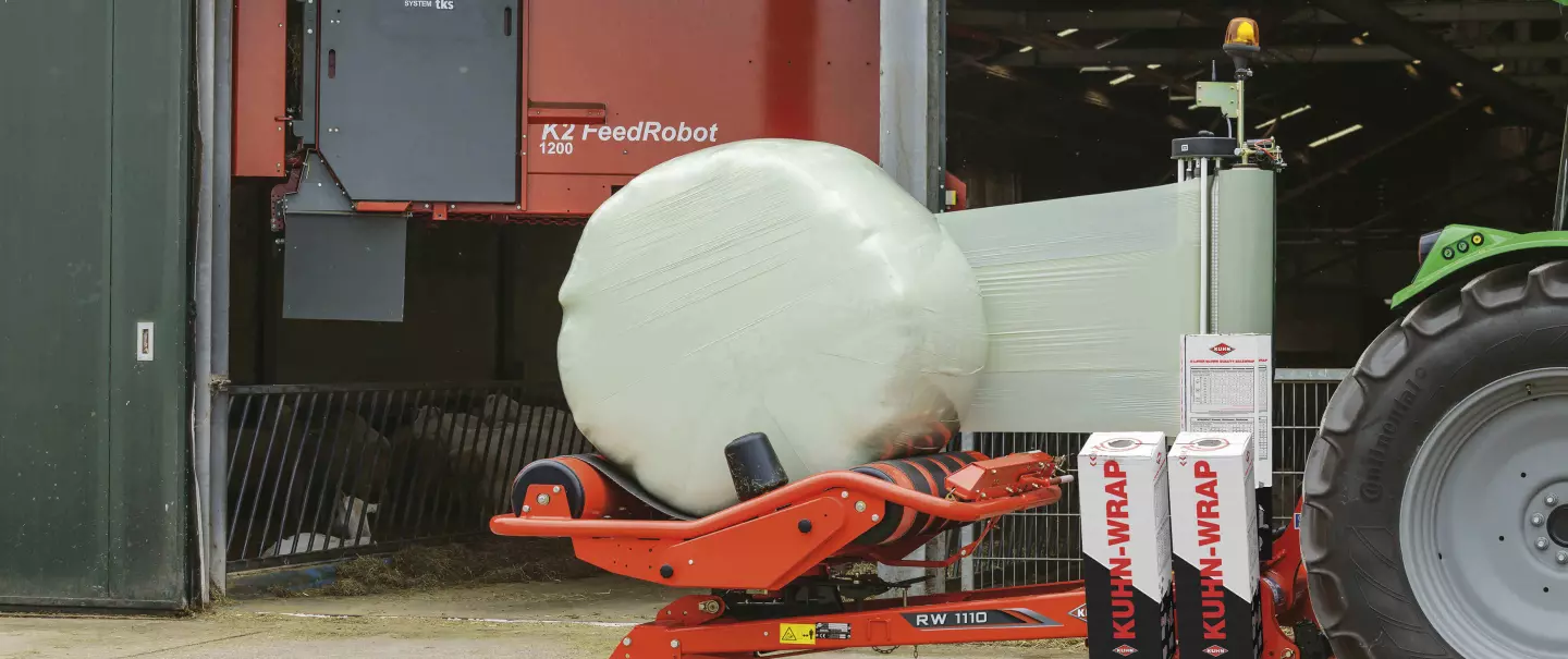 RW 1110 enrubannant une balle ronde devant une étable avec un robot d'alimentation en arrière-plan.